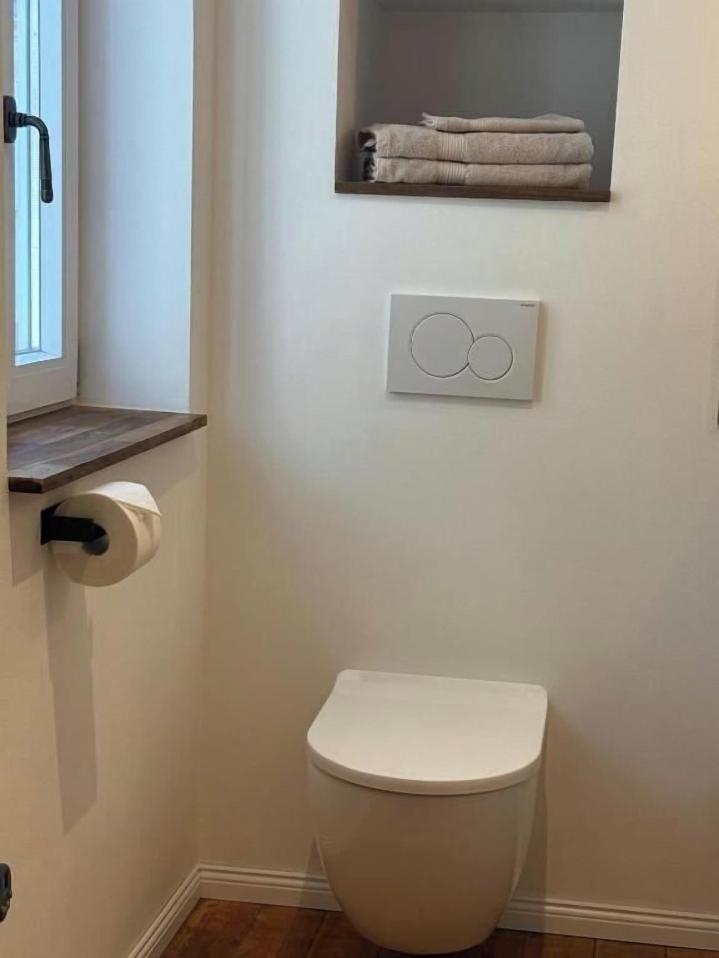 Un bagno con il water, un portaservizi con asciugamani piegati, un porta rotolo di carta igienica e un interruttore della luce fissato al muro.