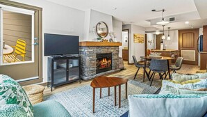 A modern living room with a stone fireplace, a flat-screen TV, and a dining area with a small island. - Contemporary 2BR w/ hot tub and Mountain Views (Dead Man`s Flats)