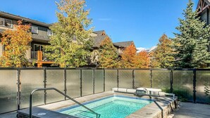 A rectangular swimming pool with a small deck area, surrounded by a metal fence and trees with autumn foliage. - Contemporary 2BR w/ hot tub and Mountain Views (Dead Man`s Flats)