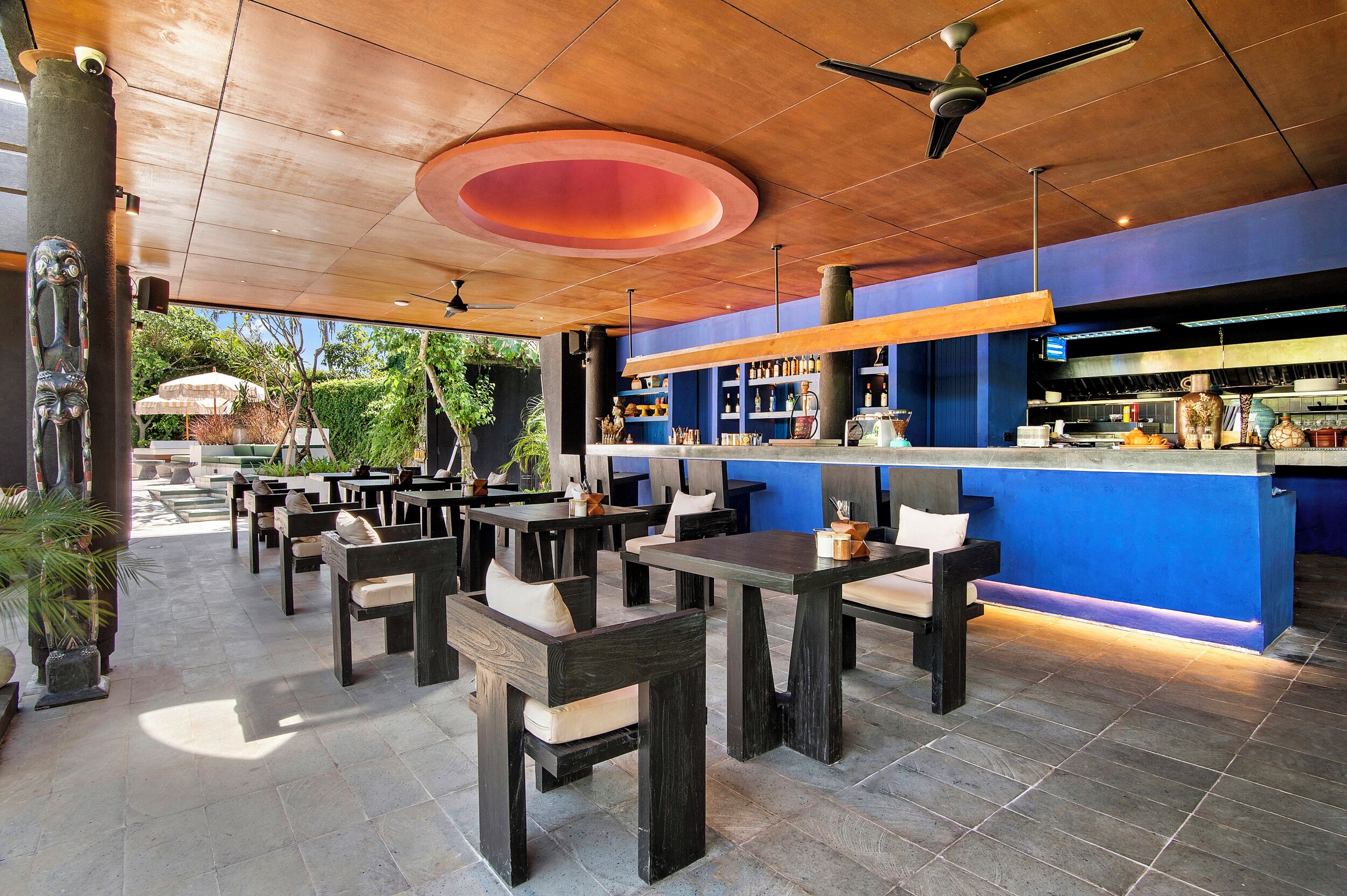An outdoor seating area with tables and chairs, a bar with shelves of bottles, and a ceiling fan.