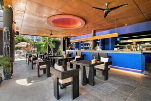 An outdoor seating area with tables and chairs, a bar with shelves of bottles, and a ceiling fan. - The Craft Hotel Ubud (Ubud)