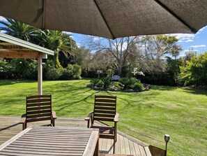 Een houten terras met twee stoelen en een tafel onder een grote parasol, met uitzicht op een verzorgd gazon met diverse planten en bomen.