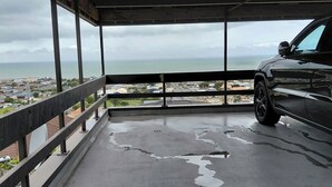 A covered outdoor area with a car parked inside, a concrete floor with some water stains, and a view of a coastal town and ocean. - Come Relax at Hilltop Nest Retreat with Ocean Views (Oamaru)