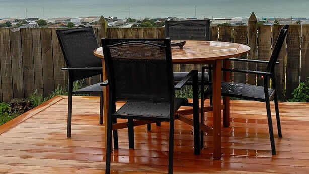 An outdoor wooden deck with a round table and four chairs, overlooking a coastal area with a wooden fence and a view of the sea. - Come Relax at Hilltop Nest Retreat with Ocean Views (Oamaru)