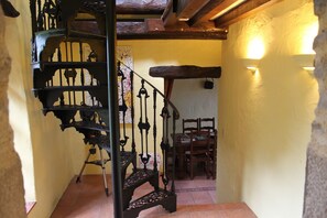 Un escalier en colimaçon orné de rampes en métal ouvragé, des chaises en bois et un luminaire fixé au mur, le tout dans une pièce aux murs jaunes et au sol carrelé.