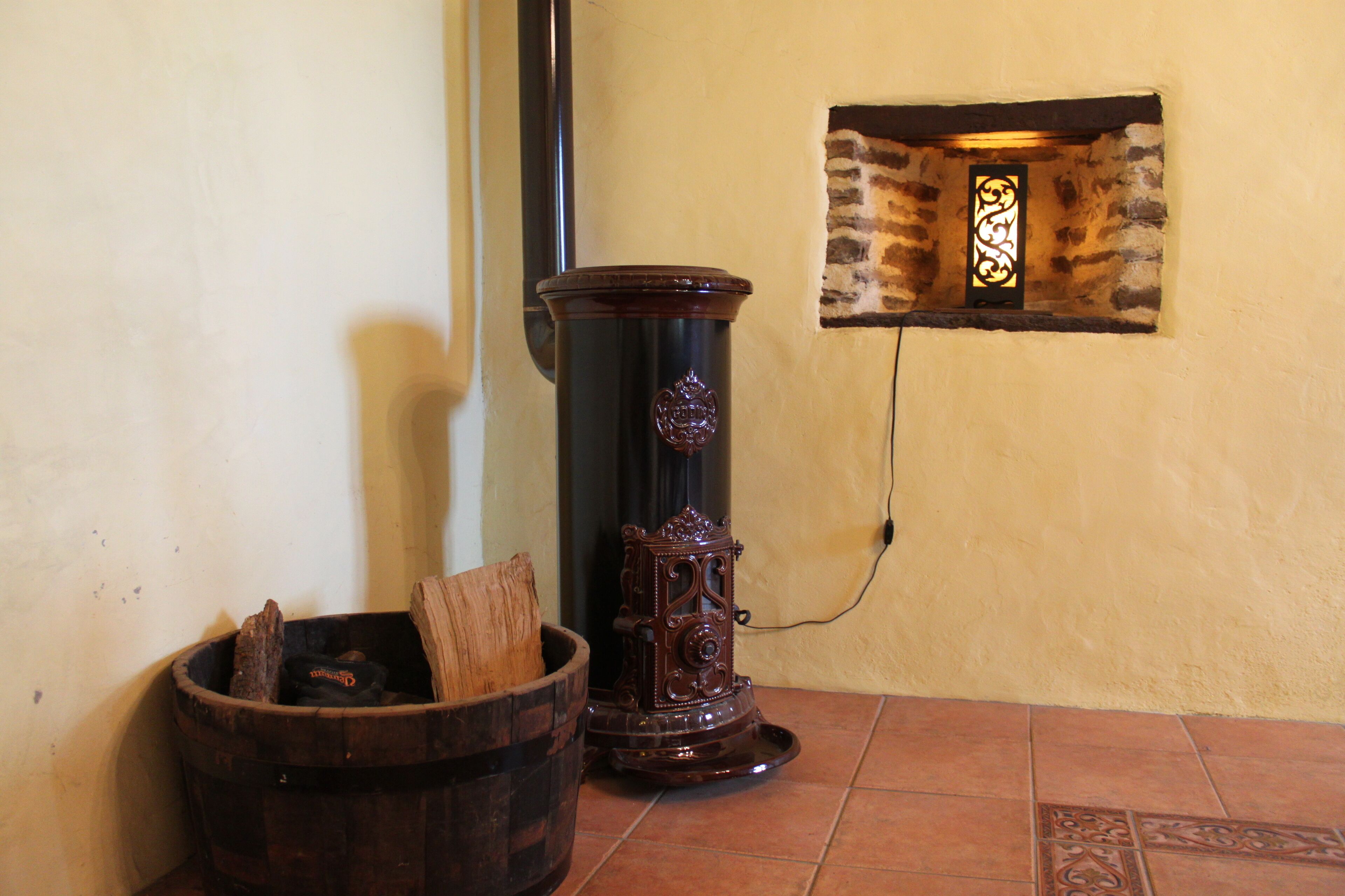 Un coin avec un seau en bois, un radiateur noir et un mur de pierre agrémenté d’une fenêtre décorative.