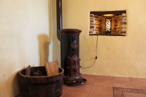 Un coin avec un seau en bois, un radiateur noir et un mur de pierre agrémenté d’une fenêtre décorative.