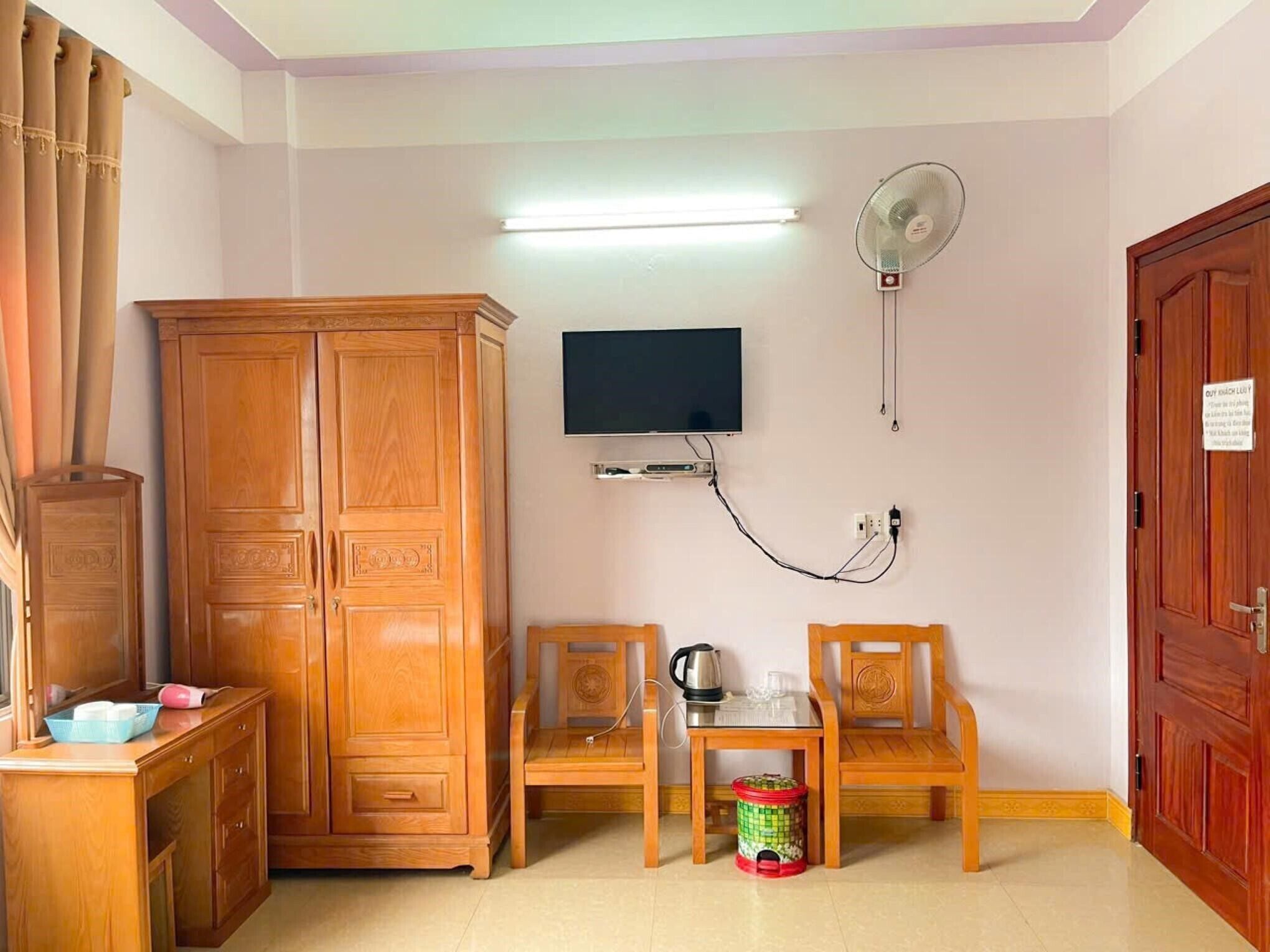 Ein Zimmer mit Holzschrank, Wand-Fernseher, Ventilator, kleinem Tisch mit Wasserkocher und Mülleimer.