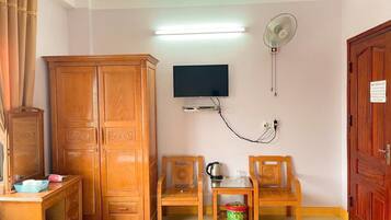Ein Zimmer mit Holzschrank, Wand-Fernseher, Ventilator, kleinem Tisch mit Wasserkocher und Mülleimer.