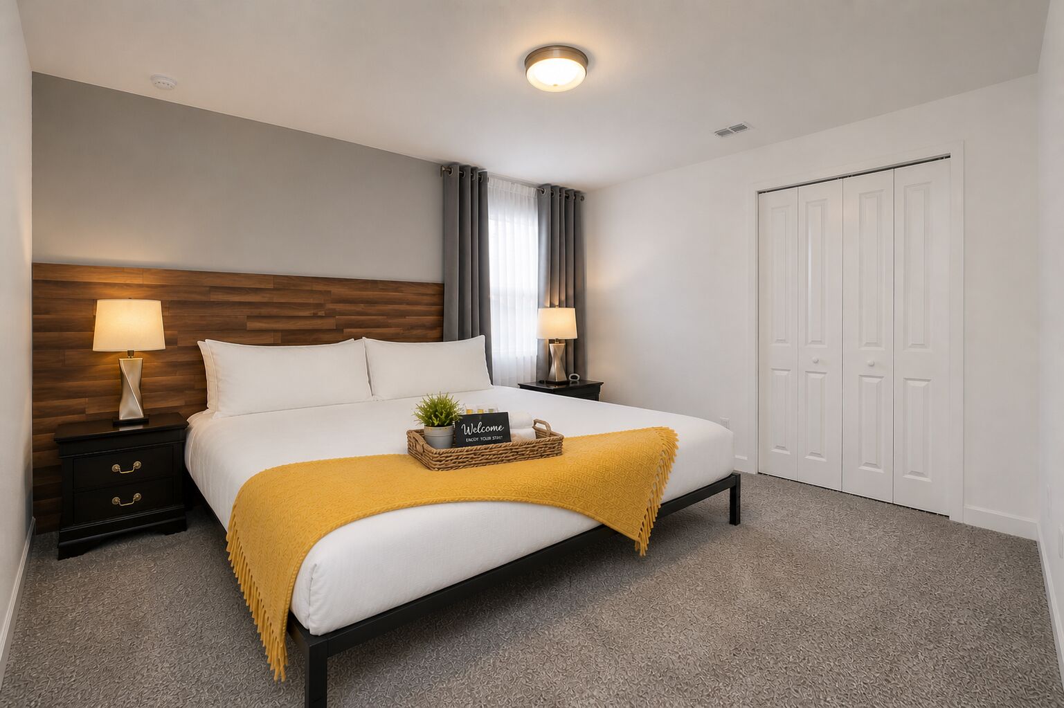 A hotel room with a bed, two bedside tables with lamps, a bedside plant, a welcome sign, and a closet.