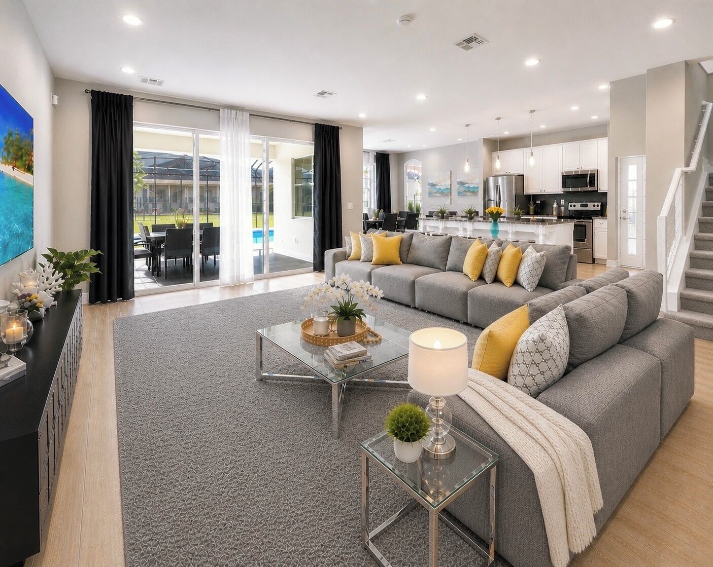 Ein modernes Wohnzimmer mit grauem Sofa, einem Glastisch und einer Küche im Hintergrund.
