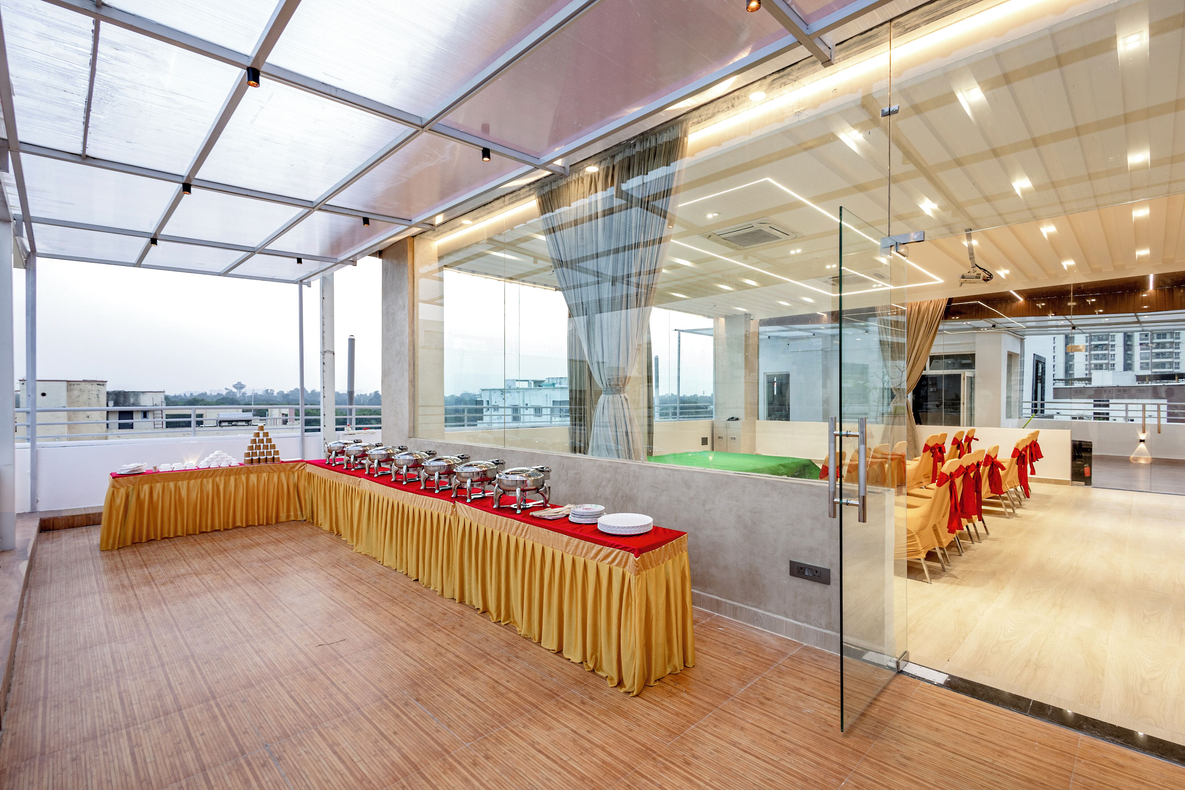 A spacious indoor event area with a long table set for a buffet, glass walls, and a view of the cityscape.