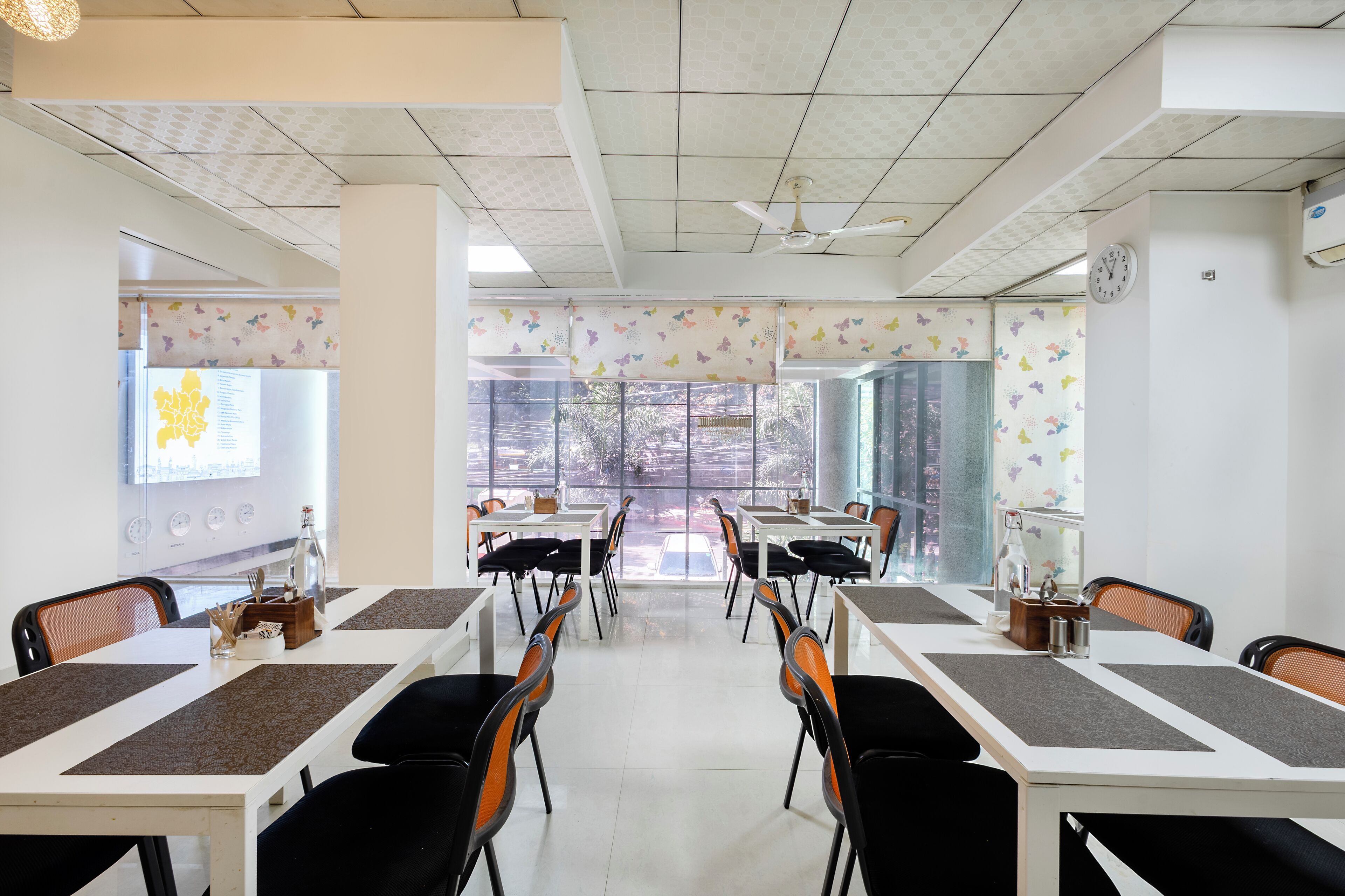 Un comedor con mesas y sillas, un ventilador de techo y ventanas grandes decoradas con mariposas.