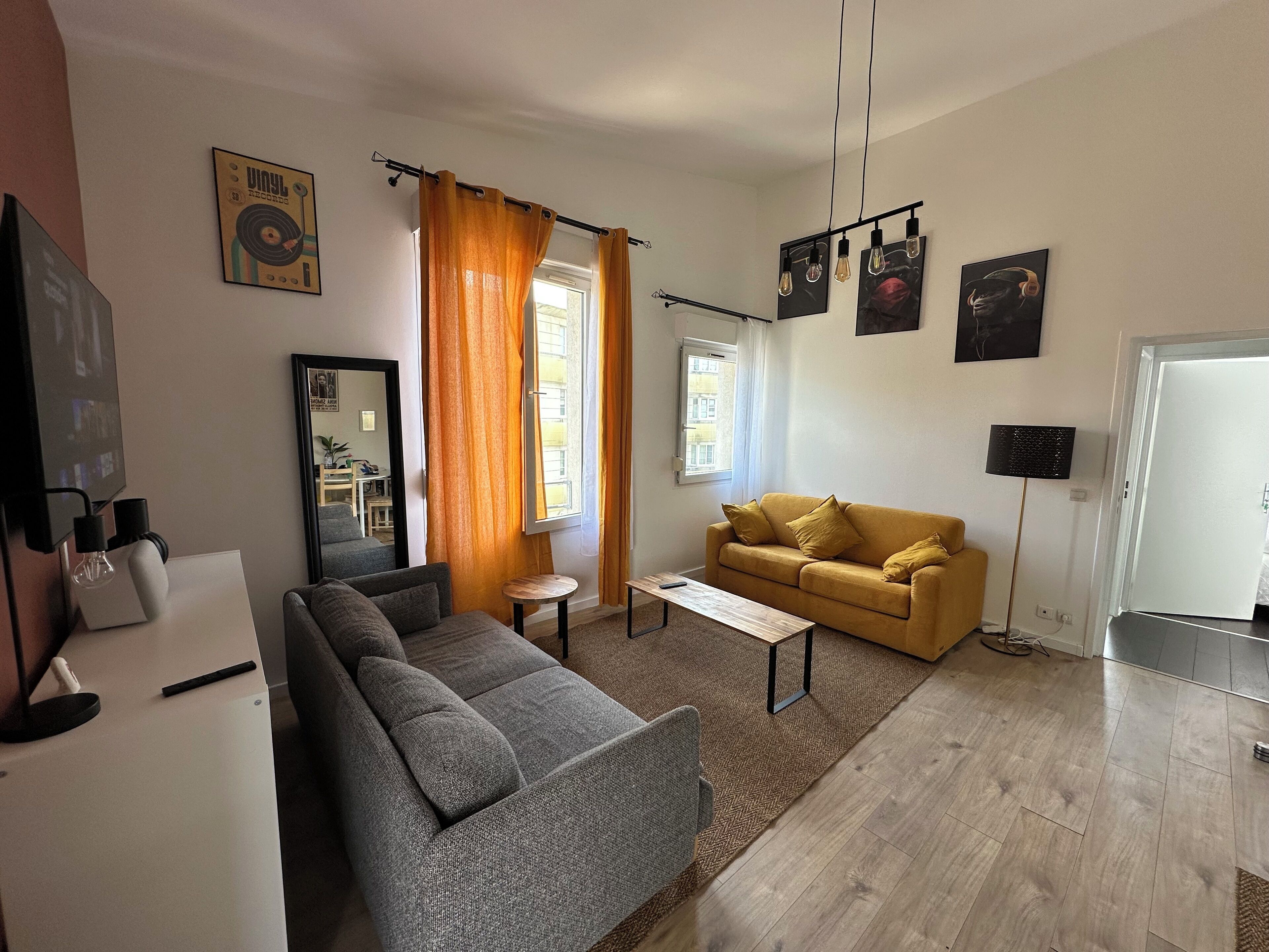 Ein Wohnzimmer mit grauem Sofa, gelbem Sofa, Couchtisch und Fernseher. Vorhänge, ein Bilderrahmen und eine Wandlampe sind ebenfalls vorhanden.