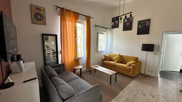 Ein Wohnzimmer mit grauem Sofa, gelbem Sofa, Couchtisch und Fernseher. Vorhänge, ein Bilderrahmen und eine Wandlampe sind ebenfalls vorhanden.
