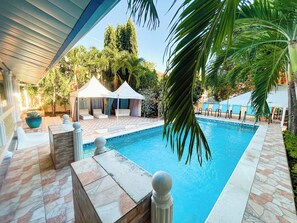 Ein Schwimmbecken mit gefliestem Sonnendeck, Liegestühlen und einer überdachten Sitzgelegenheit mit weißen Markisen.