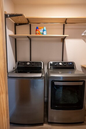 A laundry area with two washing machines and a shelf above containing Tide detergent bottles. - Modern Organic Country Home w/ Hot Tub, Pet Friendly, Near USAFA (Colorado Springs)