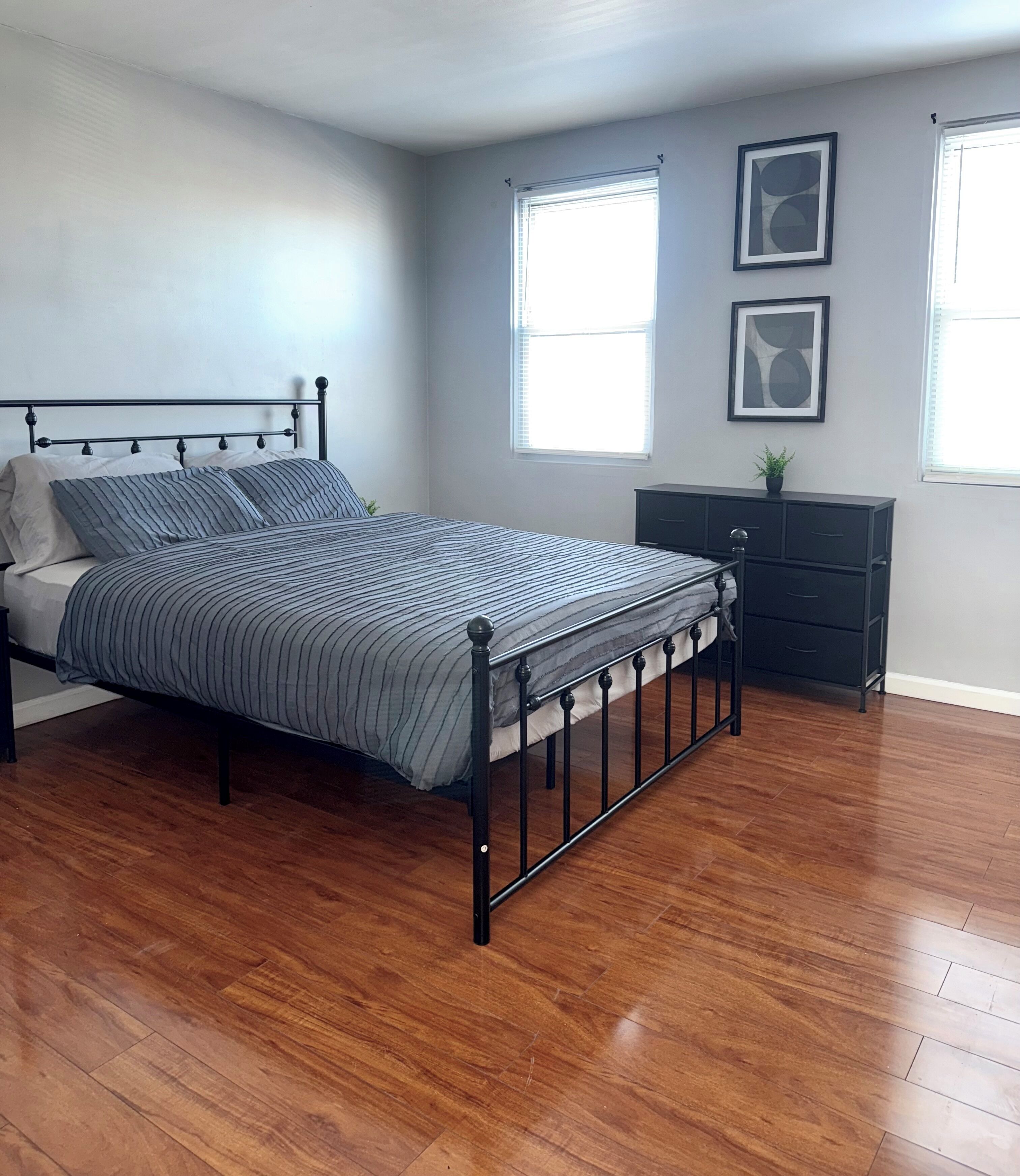 A bedroom with a bed, wooden flooring, a dresser, and two windows.
