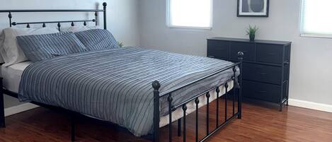 A bedroom with a bed, wooden flooring, a dresser, and two windows.