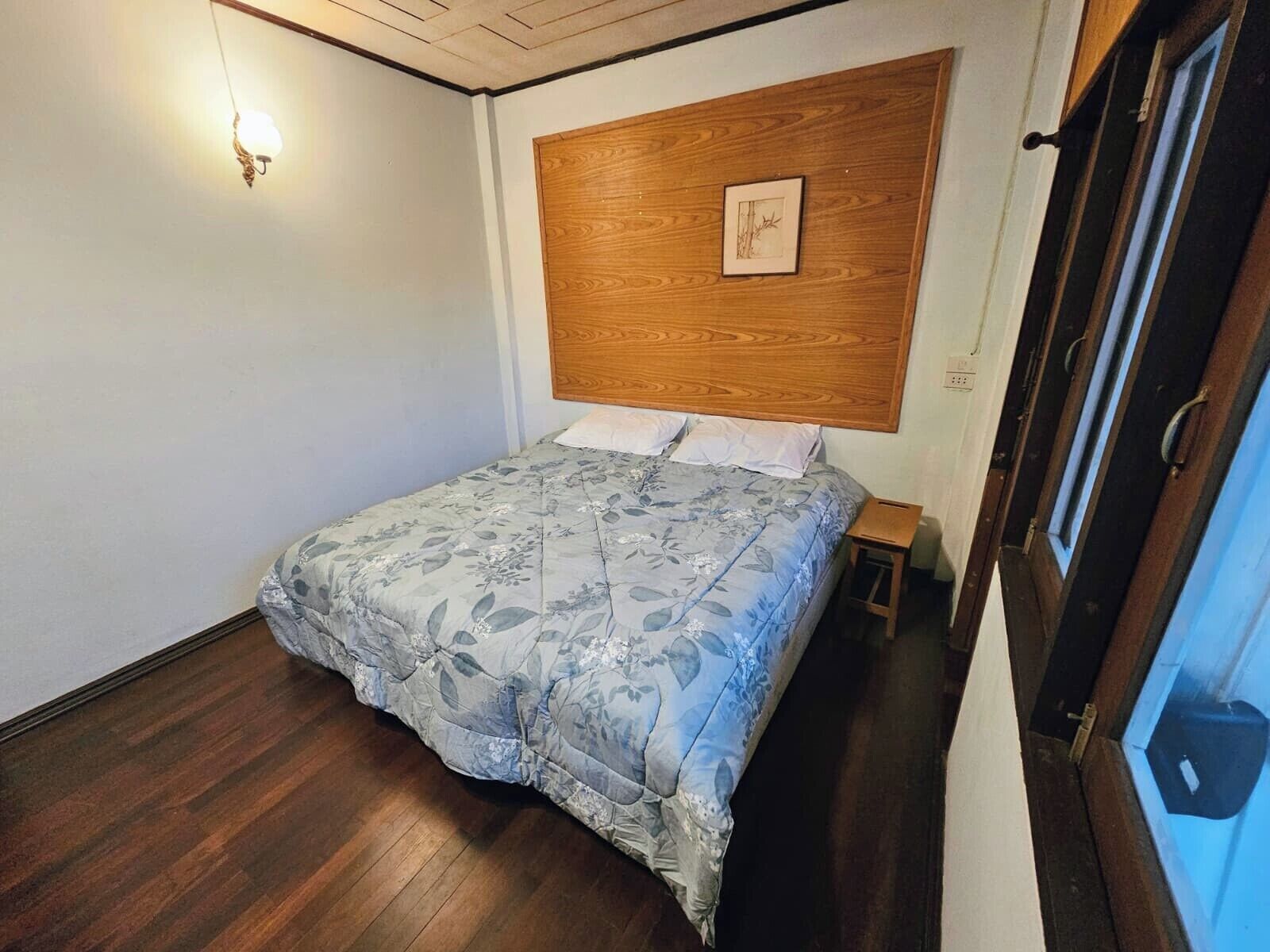 A bedroom with a wooden headboard, a bed with floral bedding, a nightstand, and a window with a view.