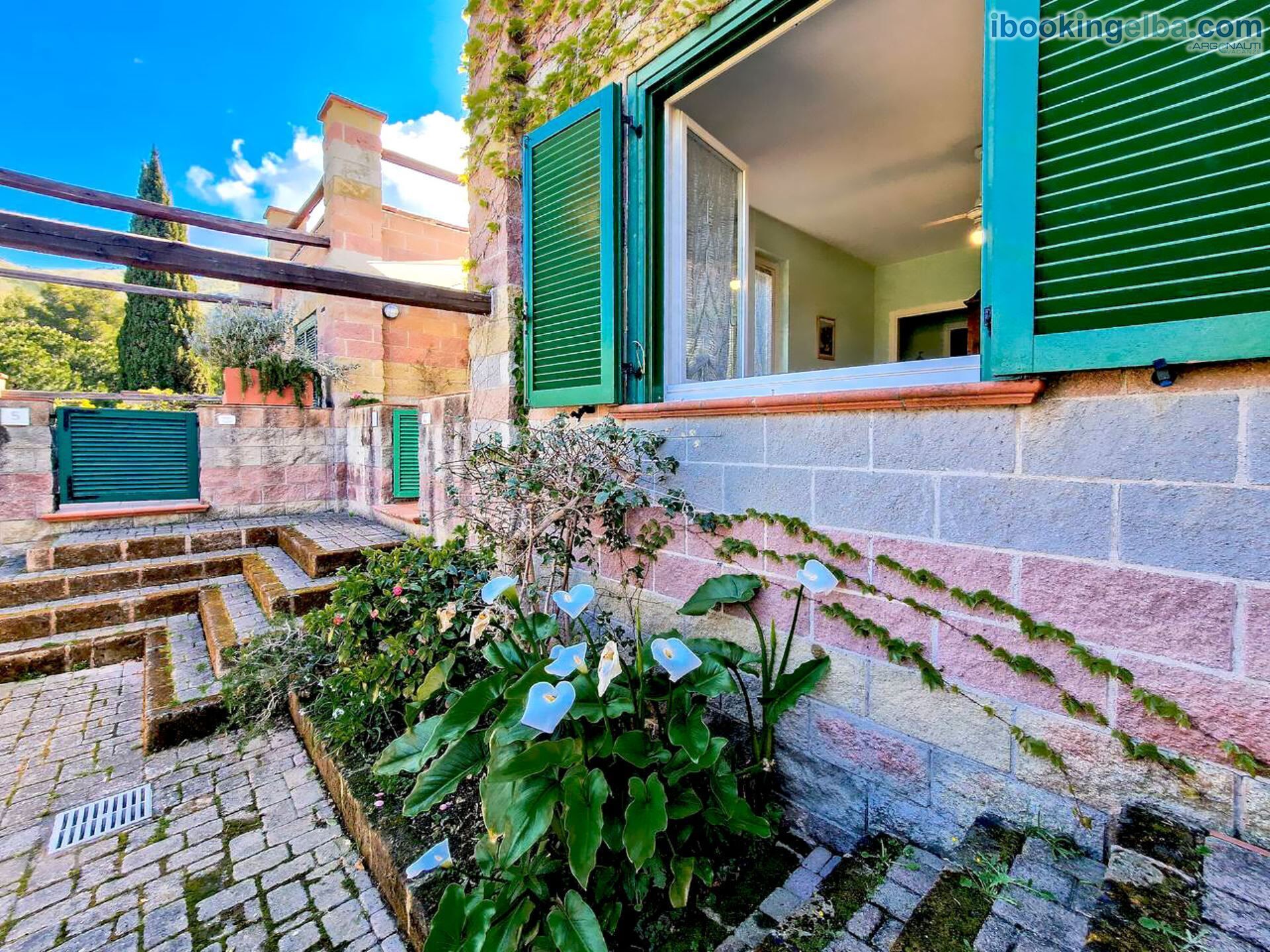Ein Innenhof mit einer Steintreppe, grünen Fensterläden und einem Garten mit herzförmigen Blüten.