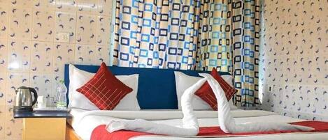 Habitación de hotel con cama, almohadas, mesita de noche, hervidor y cortina estampada.