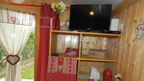 A cozy room with a TV, shelves with various items, a window with curtains, and a view of greenery outside. - CHAMBRE-GÎTE LES TOURTEREAUX EN ISERE (LE VERCORS) (Saint-Just-de-Claix)