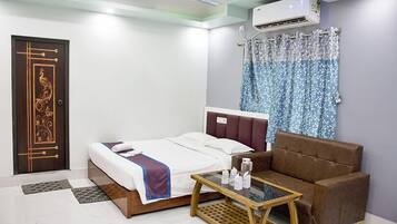 Habitación de hotel con cama, sofá marrón, mesa de centro de vidrio, cortina estampada y aire acondicionado.