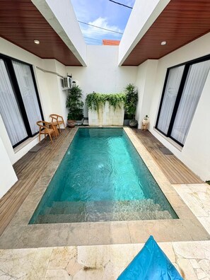 A rectangular swimming pool with a wooden deck, surrounded by white walls and large windows. - Villa Indah (Ungasan)