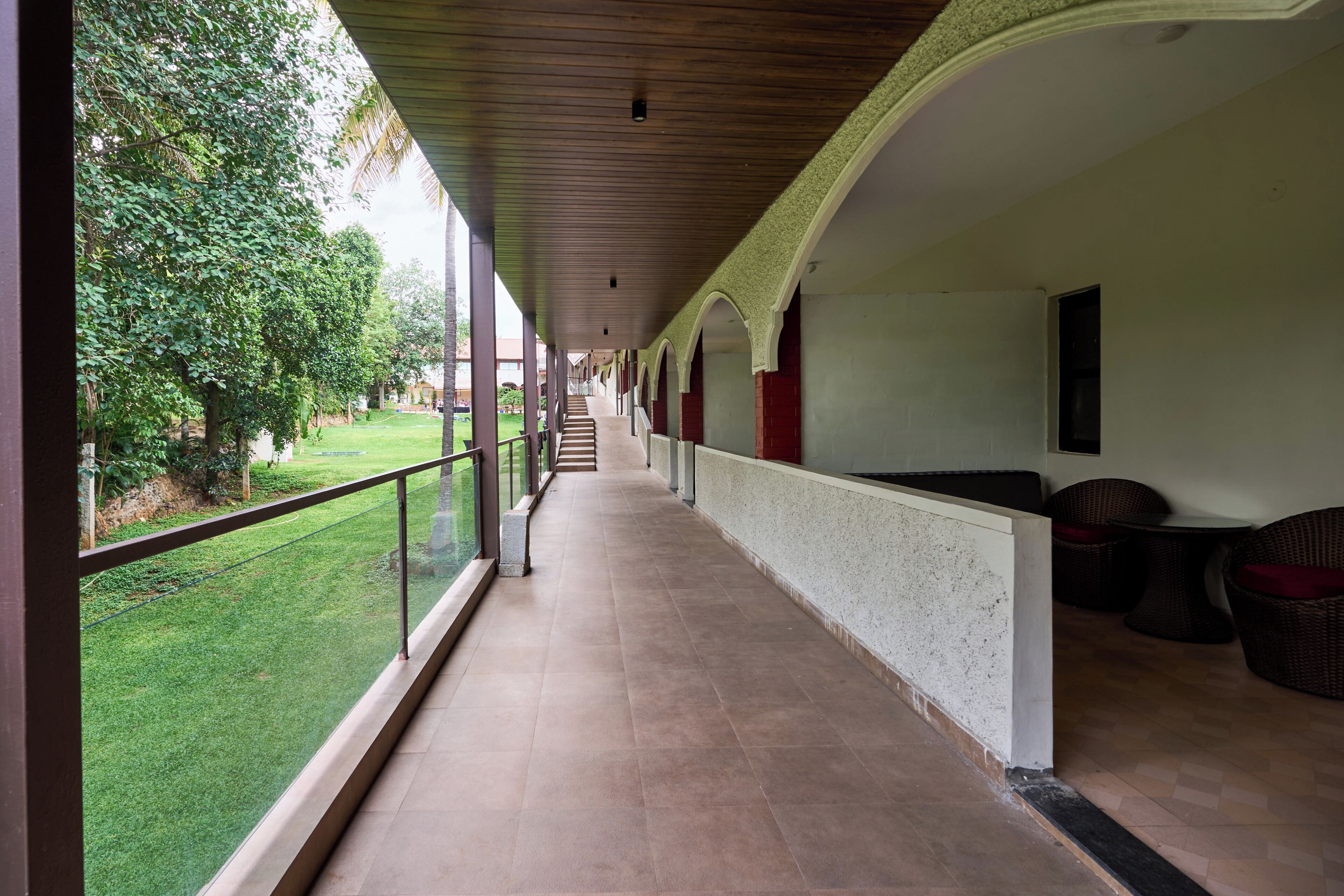 A covered corridor with a view of a green lawn and trees, leading to a building with red pillars and a tiled floor.