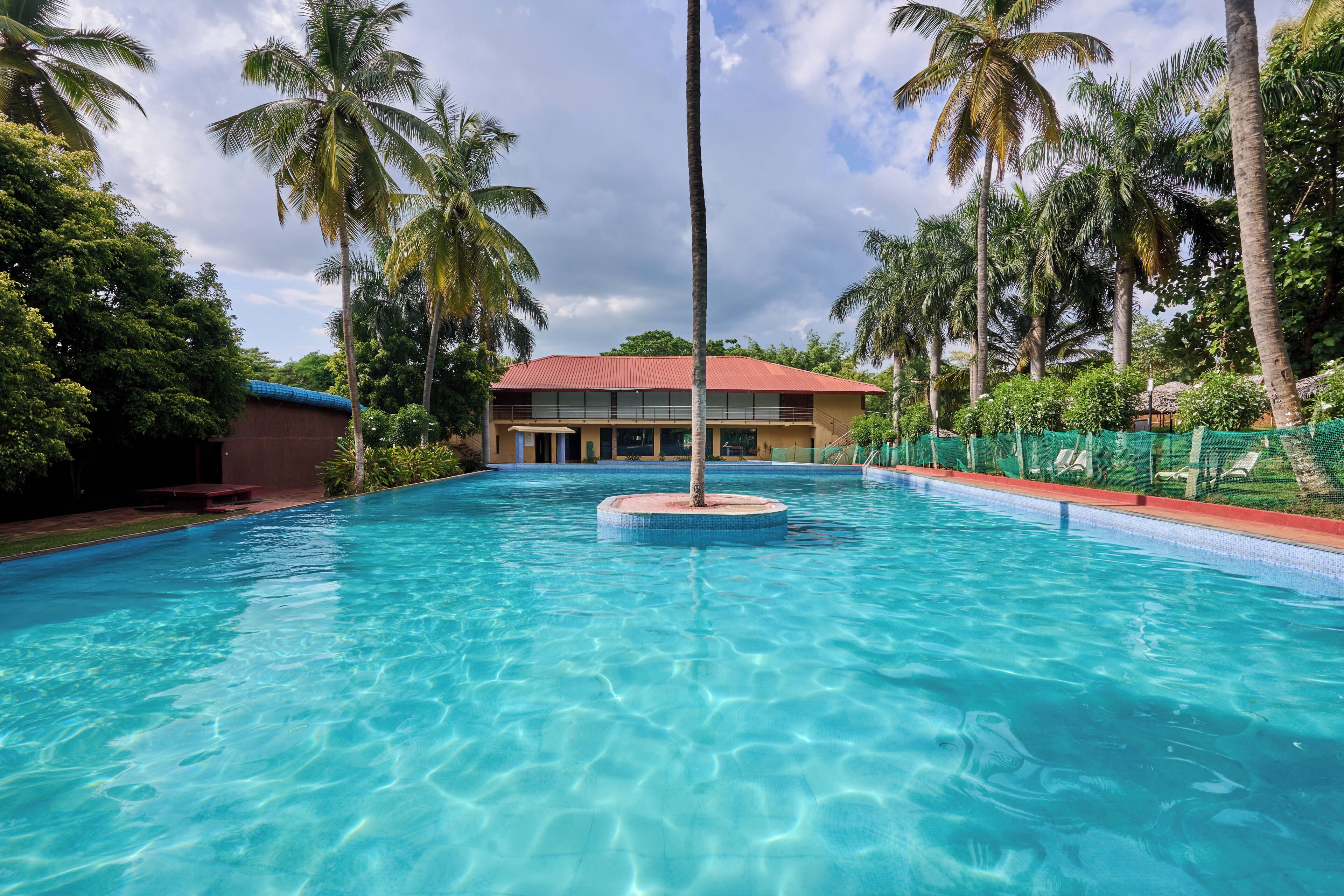 A clear swimming pool with a central island, surrounded by palm trees and a building with a red roof.