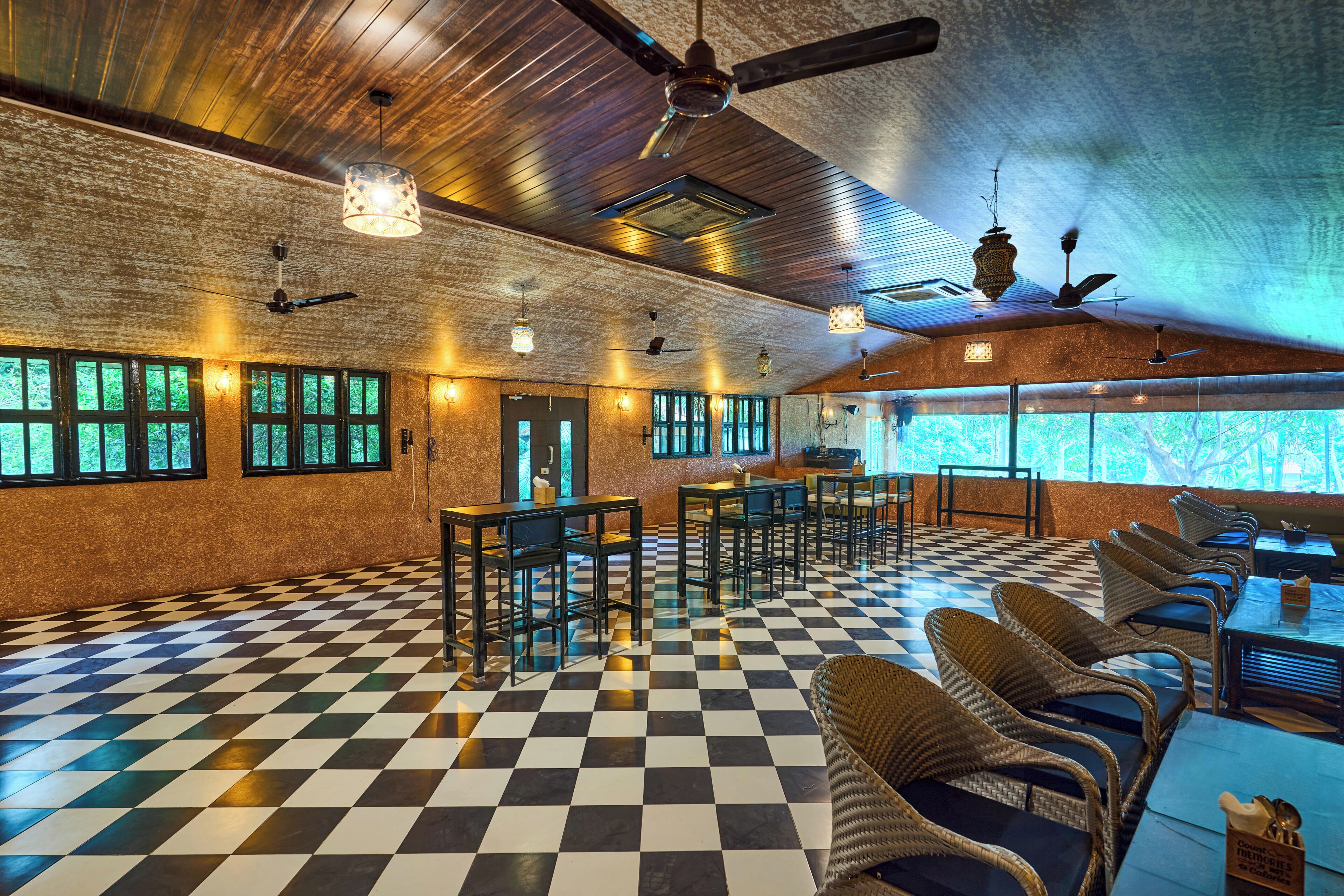 A spacious room with a checkered floor, multiple tables and chairs, ceiling fans, and large windows with a view of greenery.