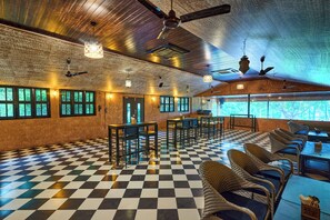 A spacious room with a checkered floor, multiple tables and chairs, ceiling fans, and large windows with a view of greenery. - Elim Resorts (Bengaluru)