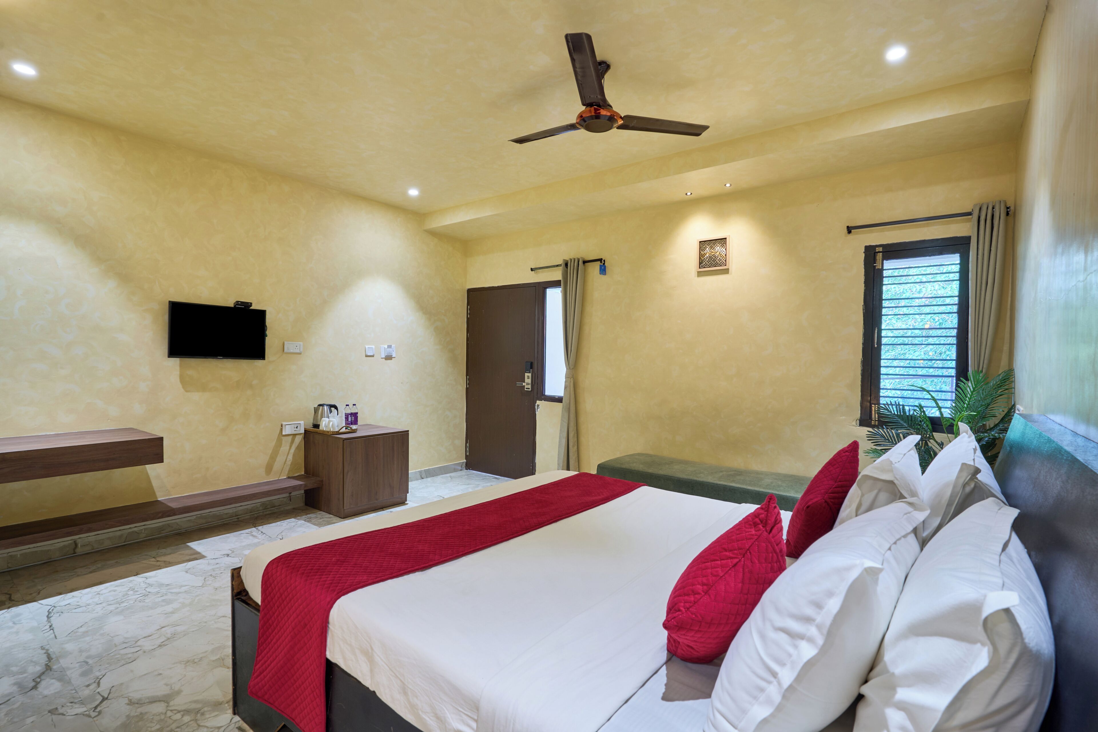 A hotel room with a bed, a TV, a ceiling fan, a window with curtains, and a small table with a kettle and bottles.