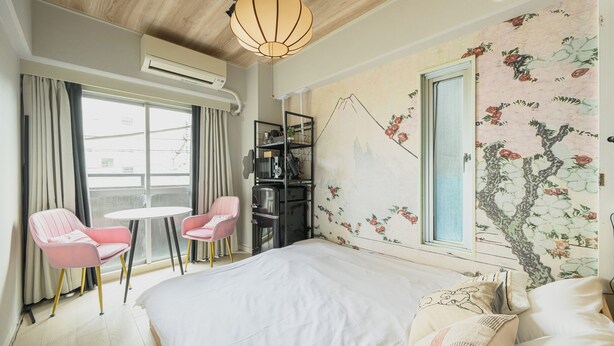 Ein Hotelzimmer mit einem Bett, zwei rosafarbenen Stühlen, einem kleinen Tisch, einem Bücherregal und einer Wand mit blumigem Wandpapier.