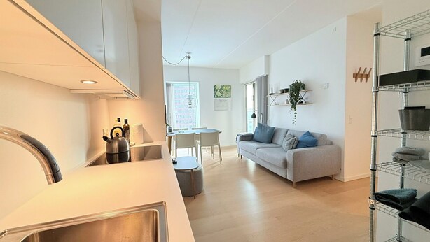 Een moderne keuken met een ingebouwde kookplaat, een eethoek met een tafel en stoelen, een grijze bank en een plank met keukengerei.