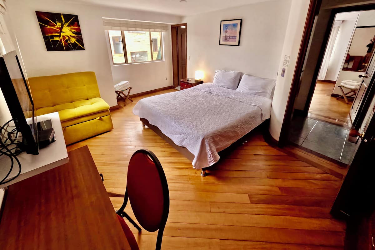 Un dormitorio con cama, sofá amarillo, escritorio con computadora y ventana con vista al exterior.