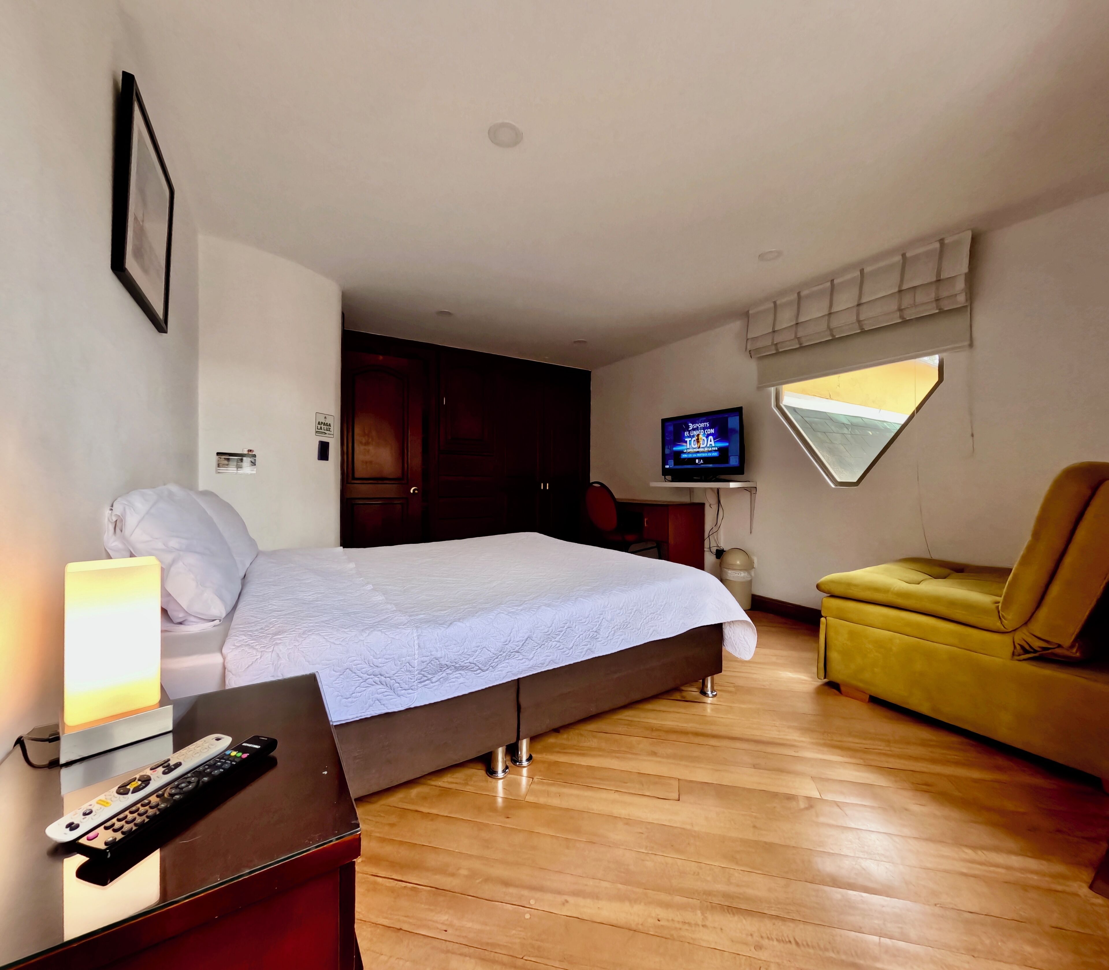 Una habitación de hotel con cama, lámpara de noche, televisión y un sillón amarillo.