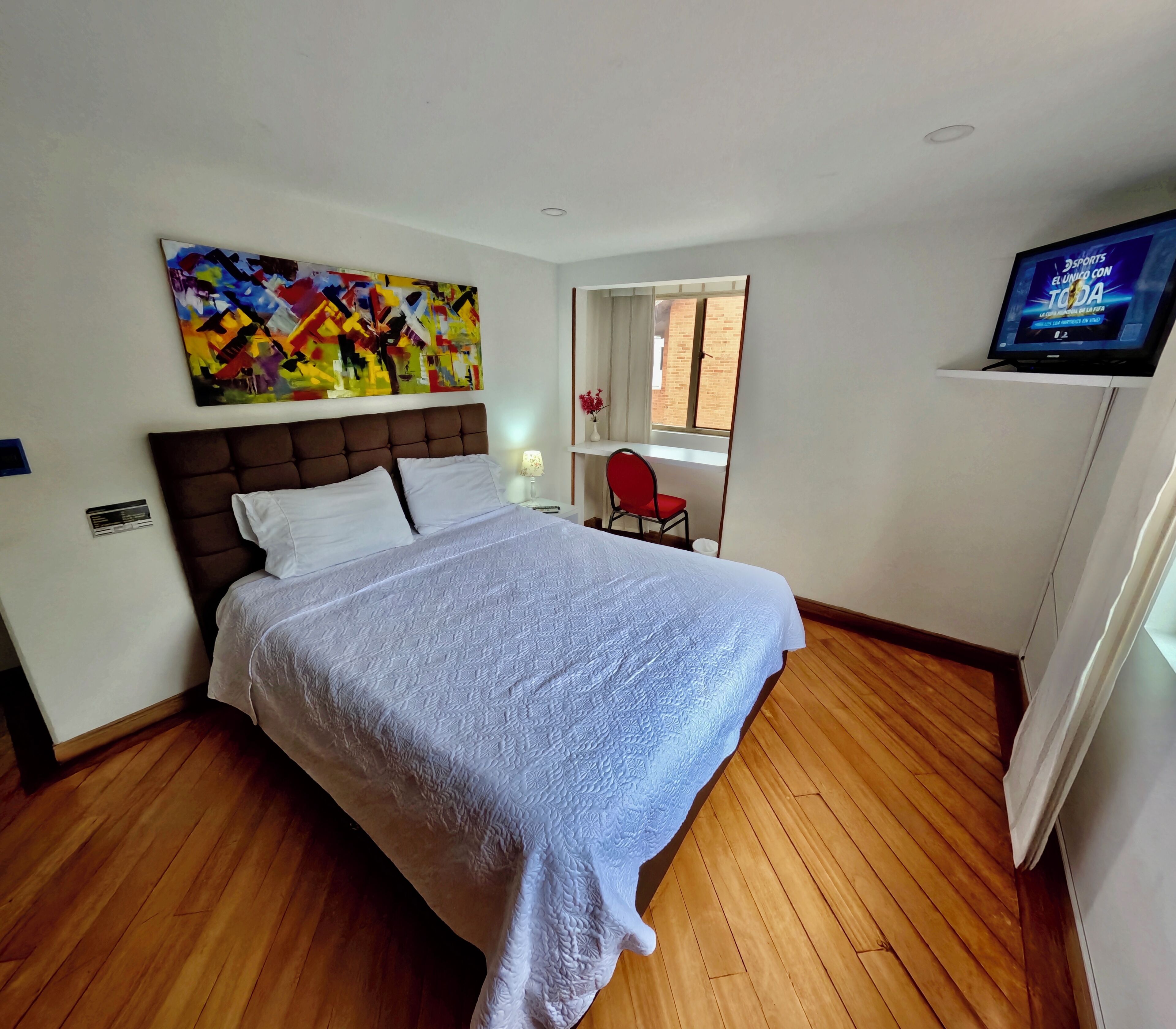 Ein Schlafzimmer mit einem großen Bett, einem Schreibtisch, einem Stuhl, einem Fernseher und einem farbenfrohen abstrakten Gemälde.