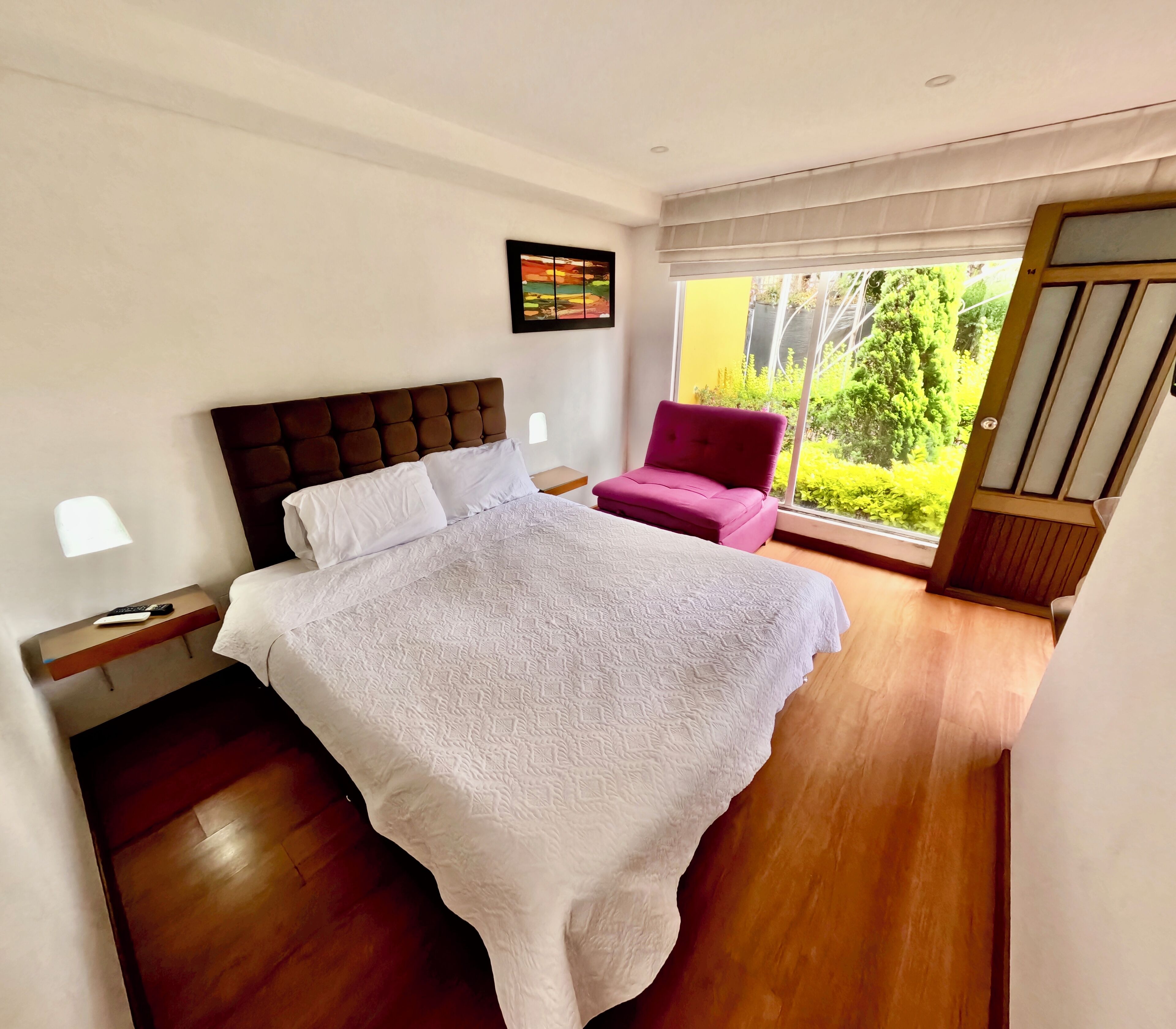 A bedroom with a large bed, a nightstand, a chair, and a window with a view of greenery.