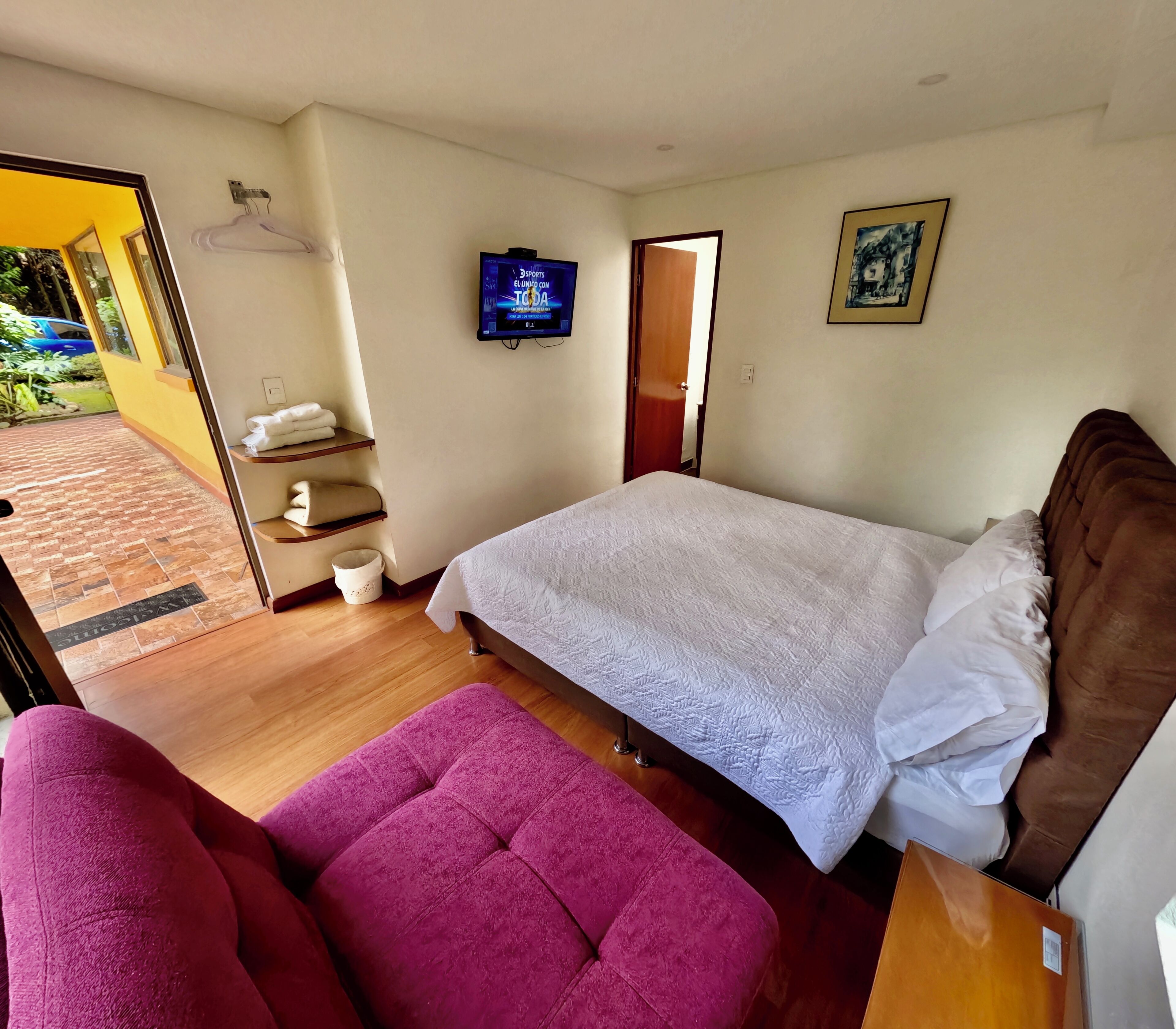 A bedroom with a bed, a TV, a wooden floor, a pink chair, and a small table.