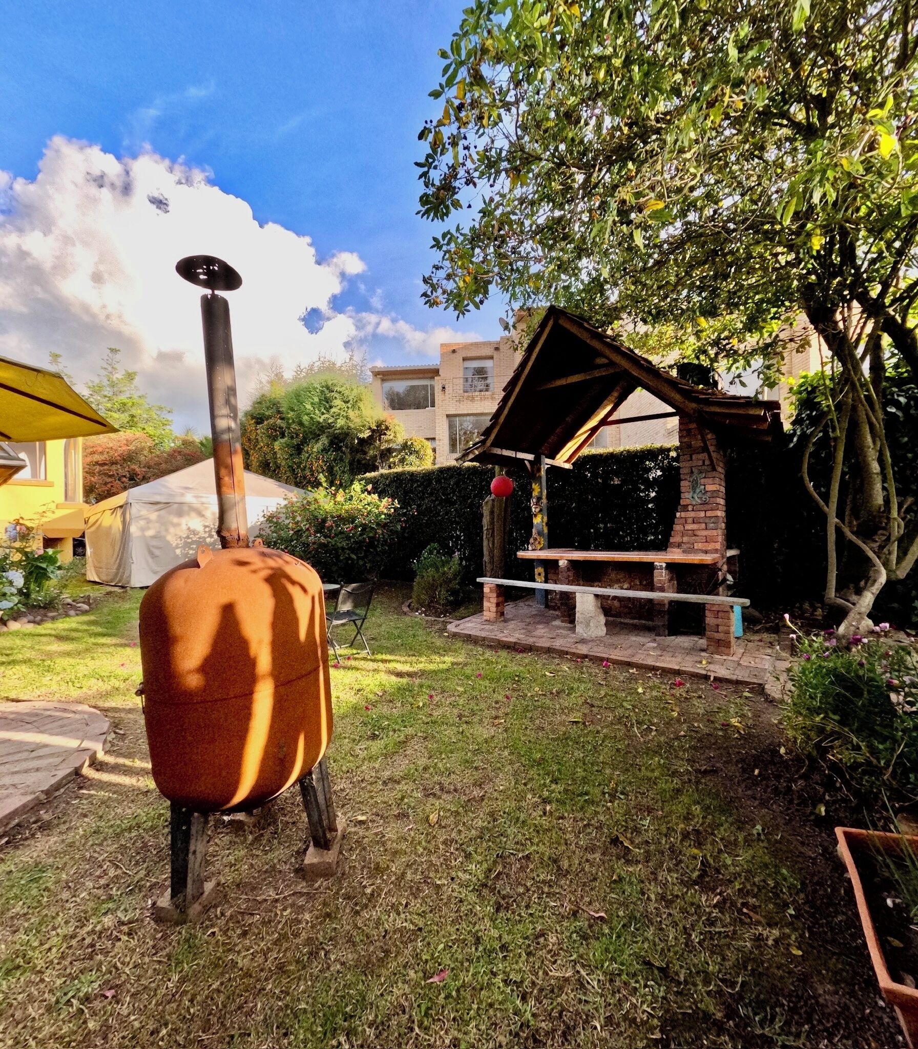 Un décor extérieur rustique, avec un grand objet cylindrique au premier plan, qui pourrait être un chauffe-eau. On aperçoit une structure en briques surmontée d’un toit, qui pourrait être un barbecue ou une cuisine d’extérieur, ainsi qu’un banc en dessous. La zone est entourée d’herbe et d’arbres, et le ciel est dégagé.