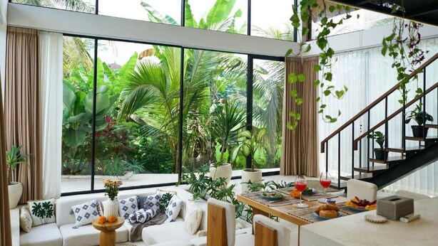 A spacious living area with a large window overlooking a lush garden, a wooden dining table with food and drinks, and a staircase with a railing. - Dzen Greenfield Villa 3 (Ubud)