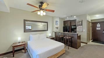 A hotel room with a bed, a desk with two chairs, a kitchenette, a ceiling fan, and a TV on the wall.