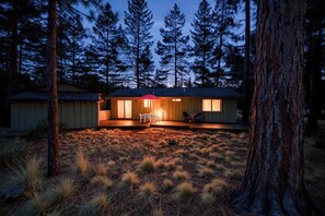 A cabin with a deck, surrounded by tall trees and a grassy area. - Cozy Sunriver Analog Escape (Sunriver)