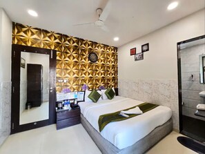 Une chambre d’hôtel comprenant un lit, une table de chevet, un miroir, un mur orné de motifs géométriques, un ventilateur de plafond et une salle de bain avec douche et lavabo.