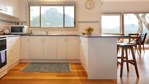 A modern kitchen with white cabinets, a built-in microwave, a large window with a mountain view, and a wooden island with bar stools. - Secluded Grampians Cottage with Mountain Views & Wildlife (Dunkeld)