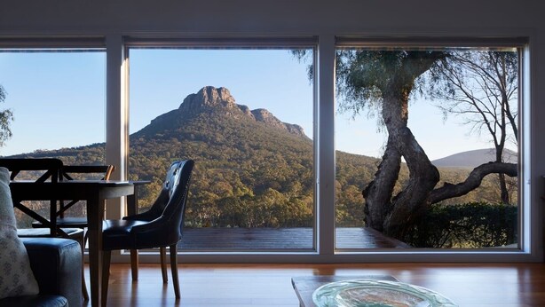 A room with a large window offering a view of a mountain and forest. There is a wooden table with a glass dish, a leather sofa, and a chair. - Secluded Grampians Cottage with Mountain Views & Wildlife (Dunkeld)