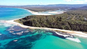 Eine Luftaufnahme einer Küstenlandschaft mit Sandstrand, klarem türkisblauem Wasser und dicht bewaldeten Hügeln.