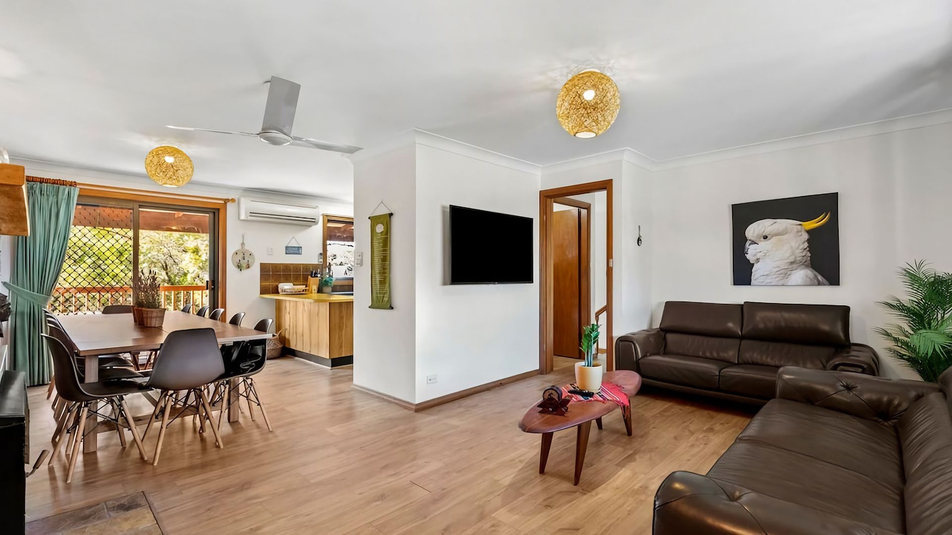 A modern living room with a brown leather sofa, a dining area with a wooden table and chairs, and a kitchen in the background.